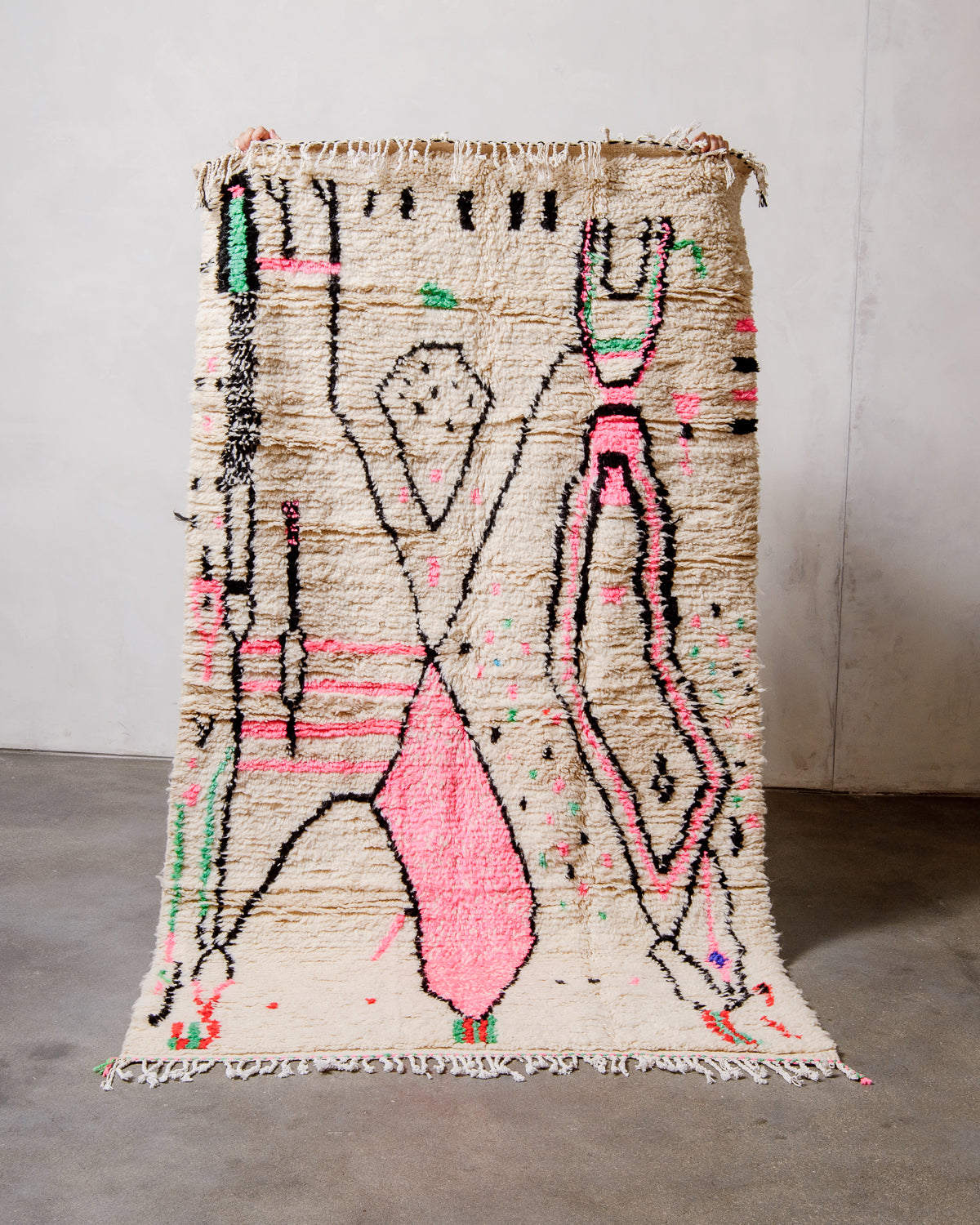 Berberlin Azilal rug made of fluffy cream wool with cotton tassels, held up against a white wall and cement floor.